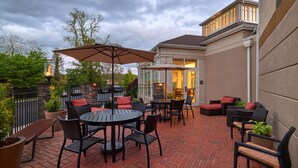 An outdoor patio with brick flooring, round tables, and black wicker chairs with red cushions.