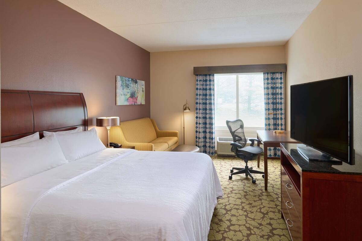 A hotel room with a large bed, a desk with a television, a chair, and a window with curtains.