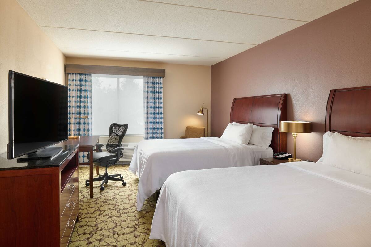 A hotel room with two beds, a desk with a television, a chair, a lamp, and a window with patterned curtains.