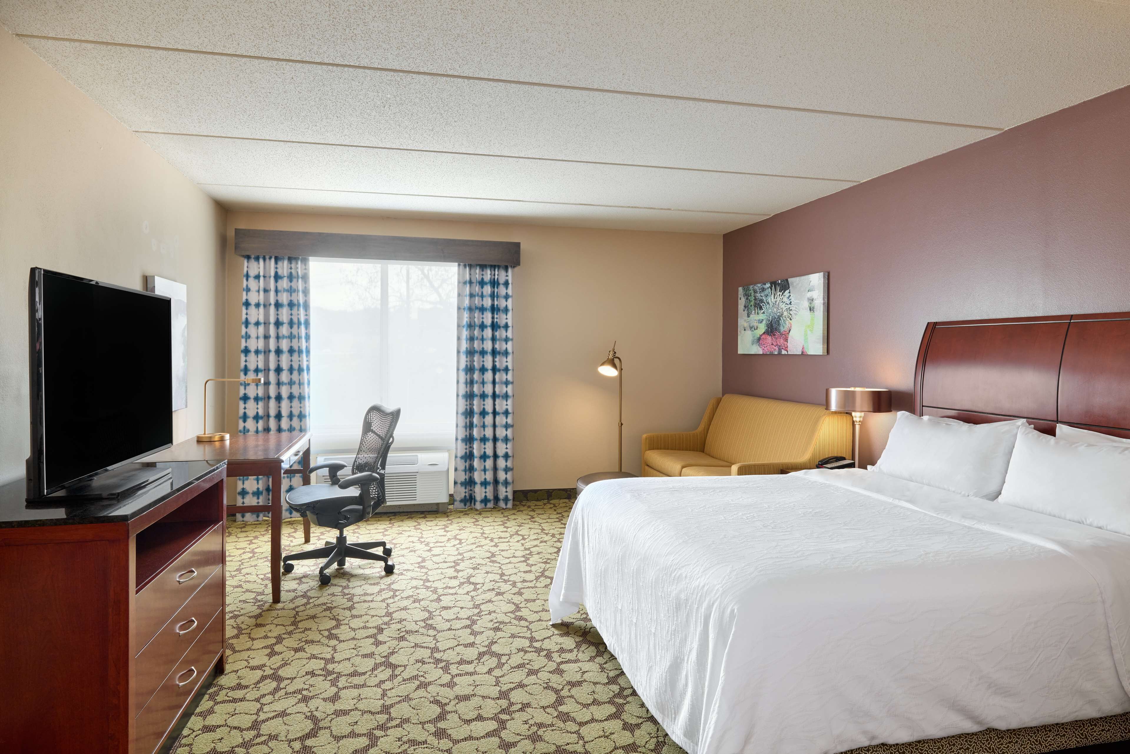 A hotel room with a bed, a desk with a television, a chair, a lamp, and a window with curtains.