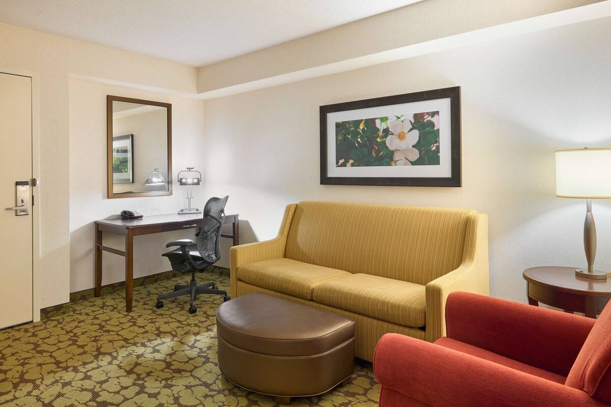 A hotel room with a sofa, a red armchair, a brown ottoman, a desk with a chair, a lamp, and a framed picture on the wall.