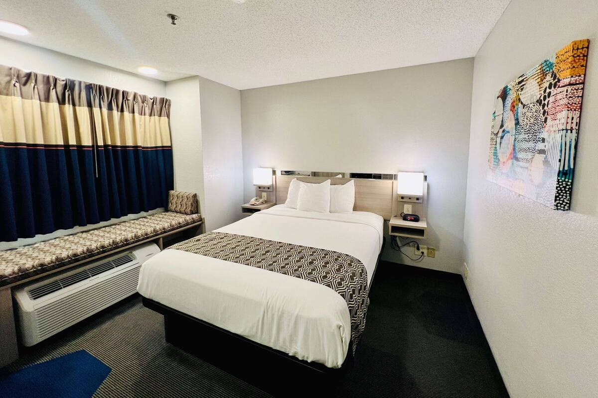 A hotel room with a large bed, bedside tables with lamps, a bench with cushions, and a wall art piece.