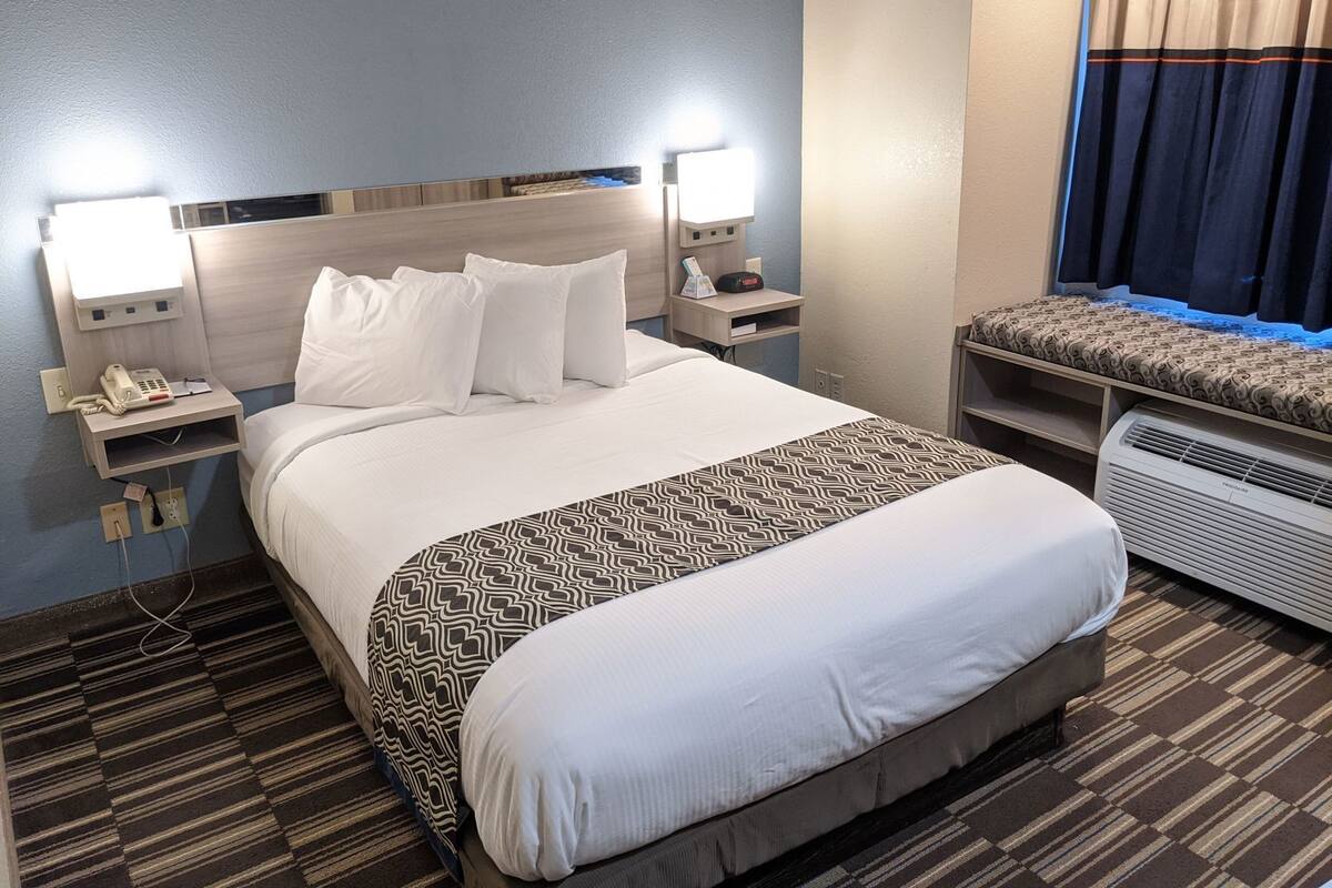 A hotel room with a large bed, bedside tables with lamps, a bedside phone, a heater, and a window with curtains.