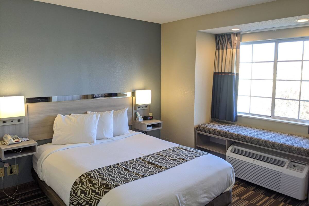 A hotel room with a large bed, bedside lamps, a heater, a window with curtains, and a small bench.
