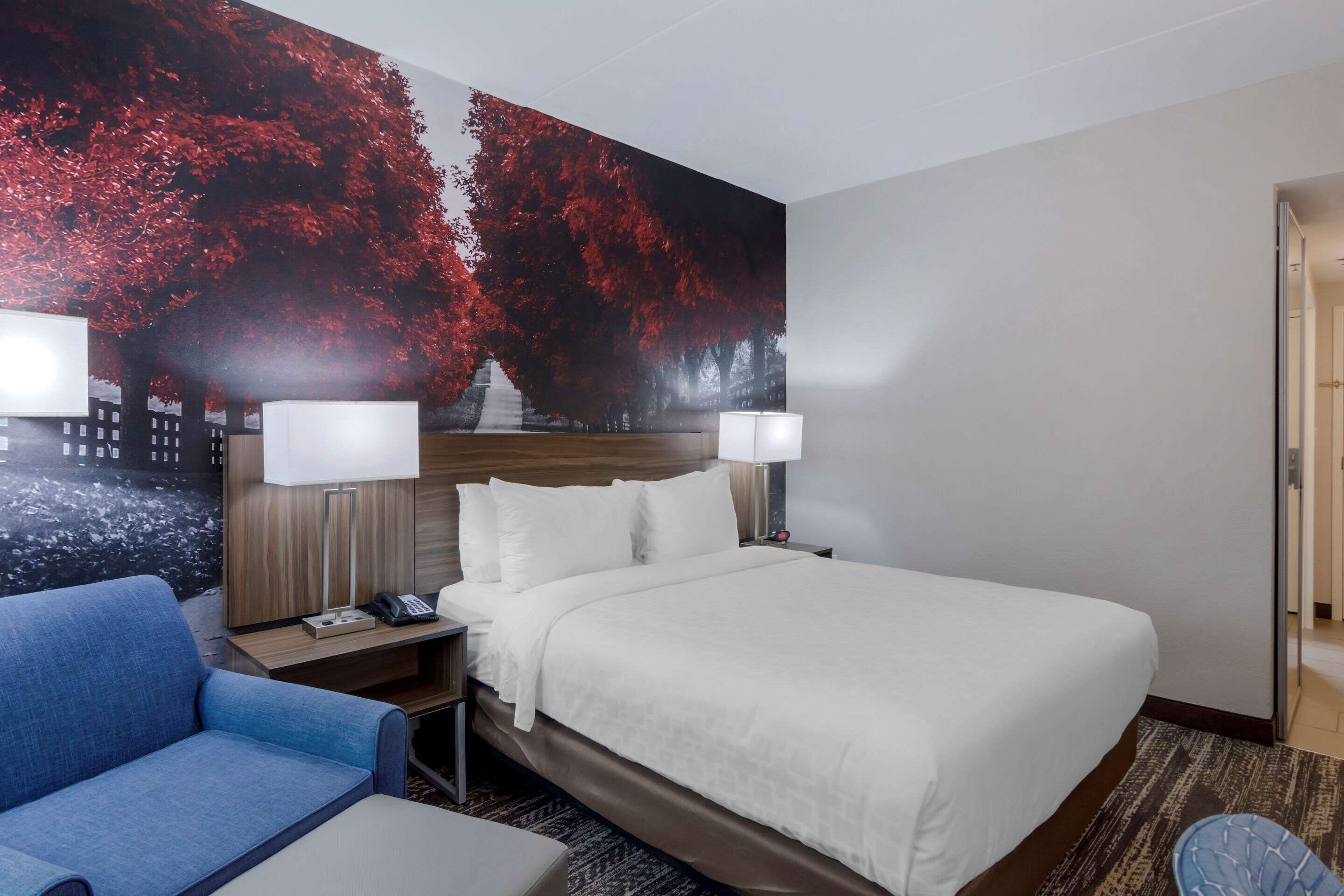Een hotelkamer met een groot bed, twee bedlampen, een nachtkastje, een blauwe fauteuil en een muurschildering van rode herfstbladeren.