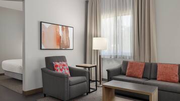 A modern hotel room with a gray sofa, a gray armchair, a small wooden table, and a floor lamp. There is a large abstract painting on the wall and a bed with a white comforter.