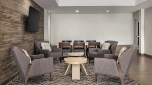 Un espace de détente moderne avec des fauteuils gris à revêtement en tissu, une table ronde centrale et un téléviseur à écran plat fixé à un mur en bois.