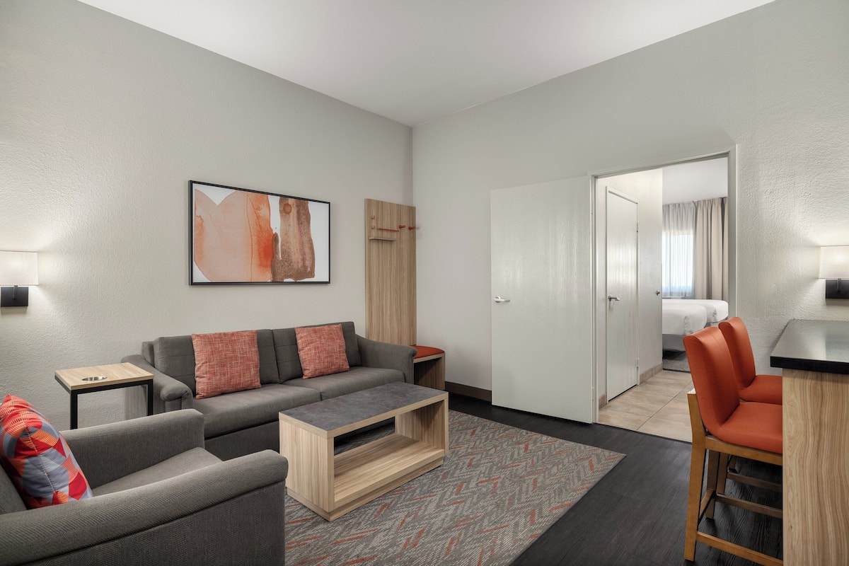 Een moderne woonkamer met een grijze bank, een houten salontafel en een rode fauteuil. Aan de muur hangt een schilderij en er is een deur die naar een slaapkamer leidt.