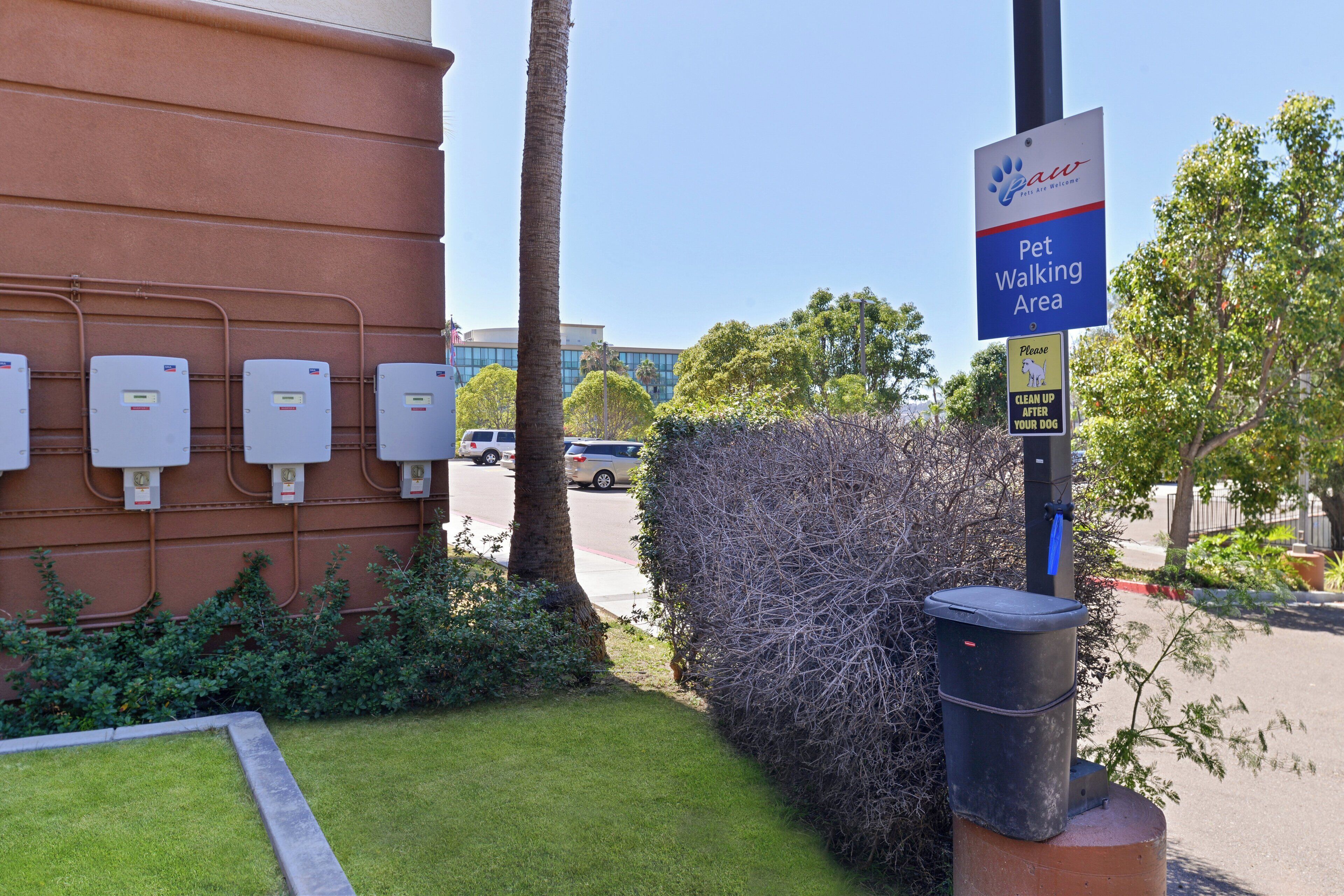 Zona donde se admiten mascotas, con un cartel que indica que es una zona para pasear perros, un cubo de basura y un edificio con contadores.