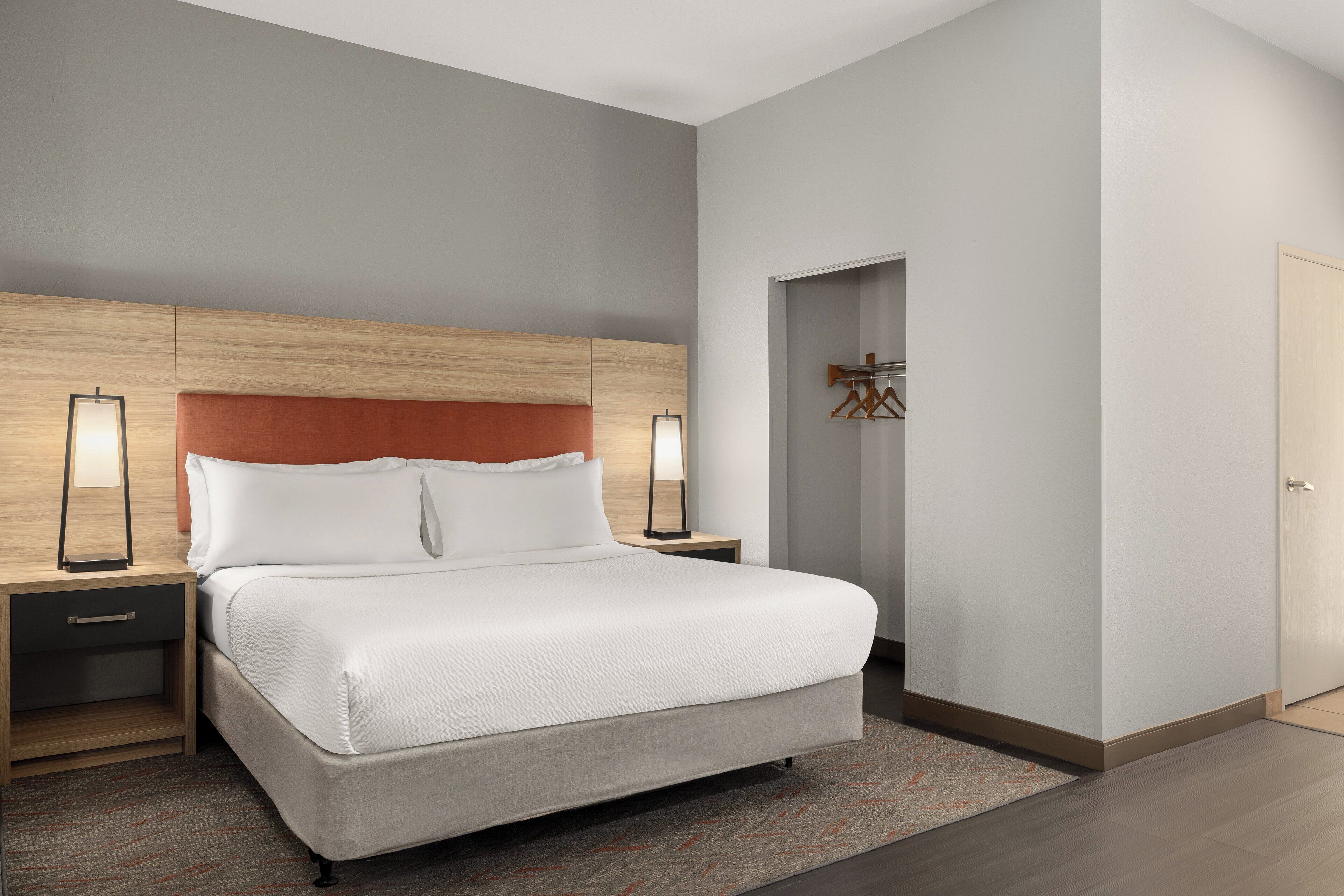 A modern hotel room with a large bed, wooden headboard, two bedside tables with lamps, a small walk-in closet, and a door to the right.