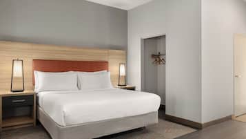 A modern hotel room with a large bed, wooden headboard, two bedside tables with lamps, a small walk-in closet, and a door to the right.