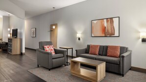 Un salon moderne avec un canapé gris, un fauteuil et une table basse. Un tableau orne le mur et un téléviseur est visible à l’arrière-plan.