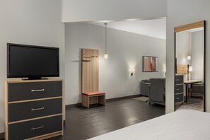 Une chambre d’hôtel moderne équipée d’un téléviseur à écran plat posé sur un meuble en bois, d’un petit banc en bois, d’un bureau avec une chaise et d’un grand miroir.