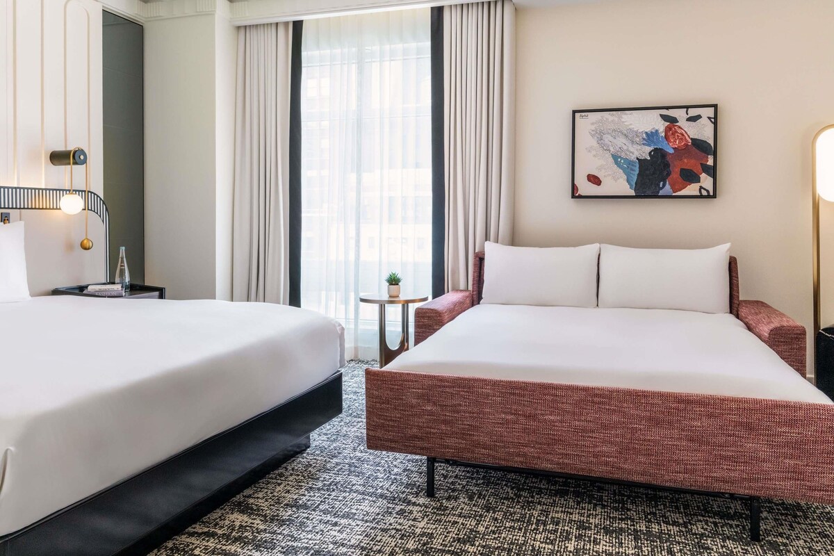 A hotel room with two beds, a small table with a plant, a painting on the wall, and a window with curtains.