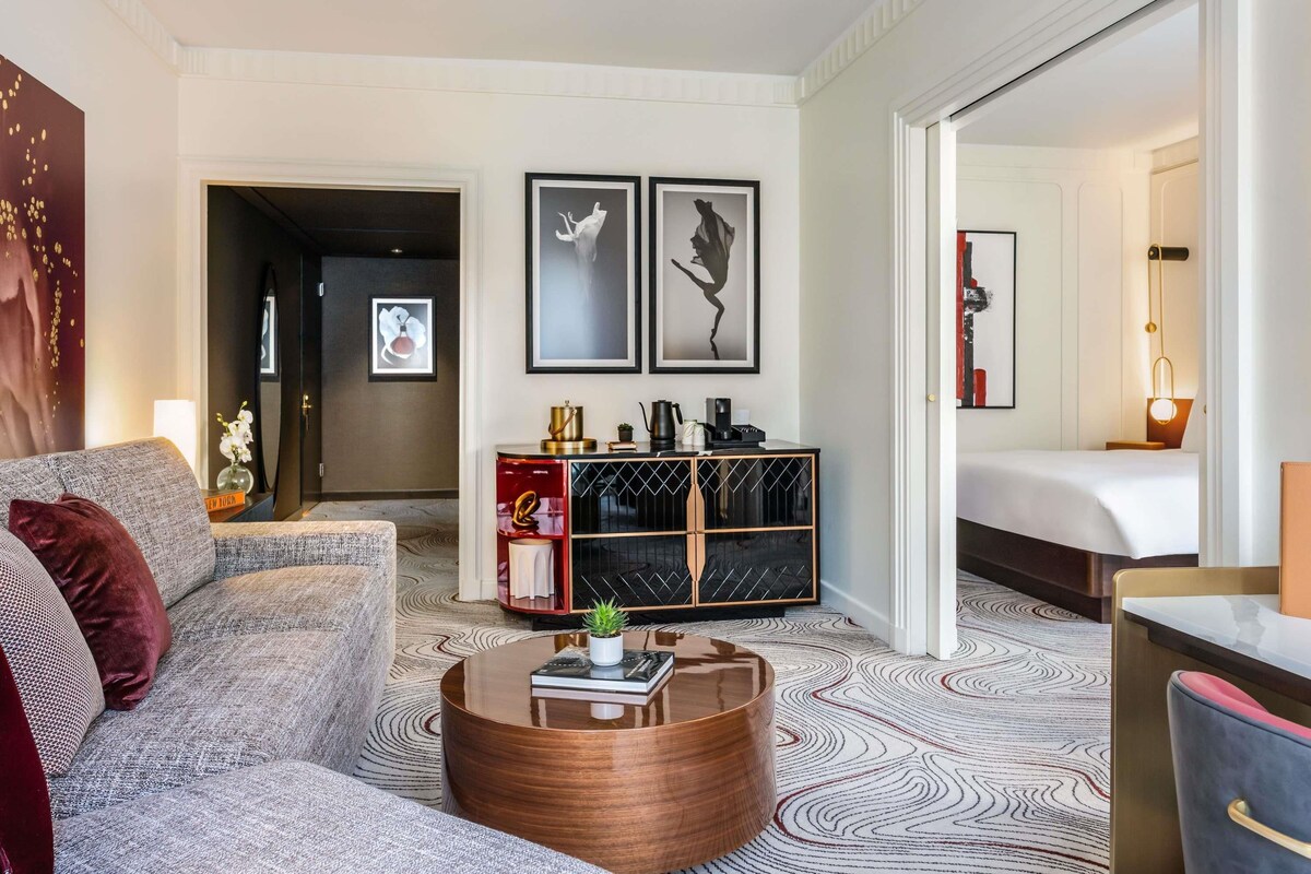 A modern hotel room with a sofa, a round coffee table, a side table with a lamp, a bed, and a console table with decorative items.
