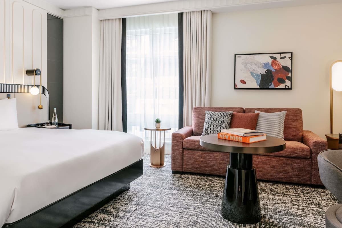 A hotel room with a bed, a sofa, a small table, a potted plant, and a painting on the wall.