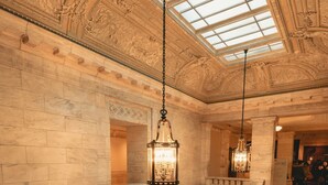 A grand interior with ornate stone walls, a glass-paneled ceiling, and a staircase with a balustrade.