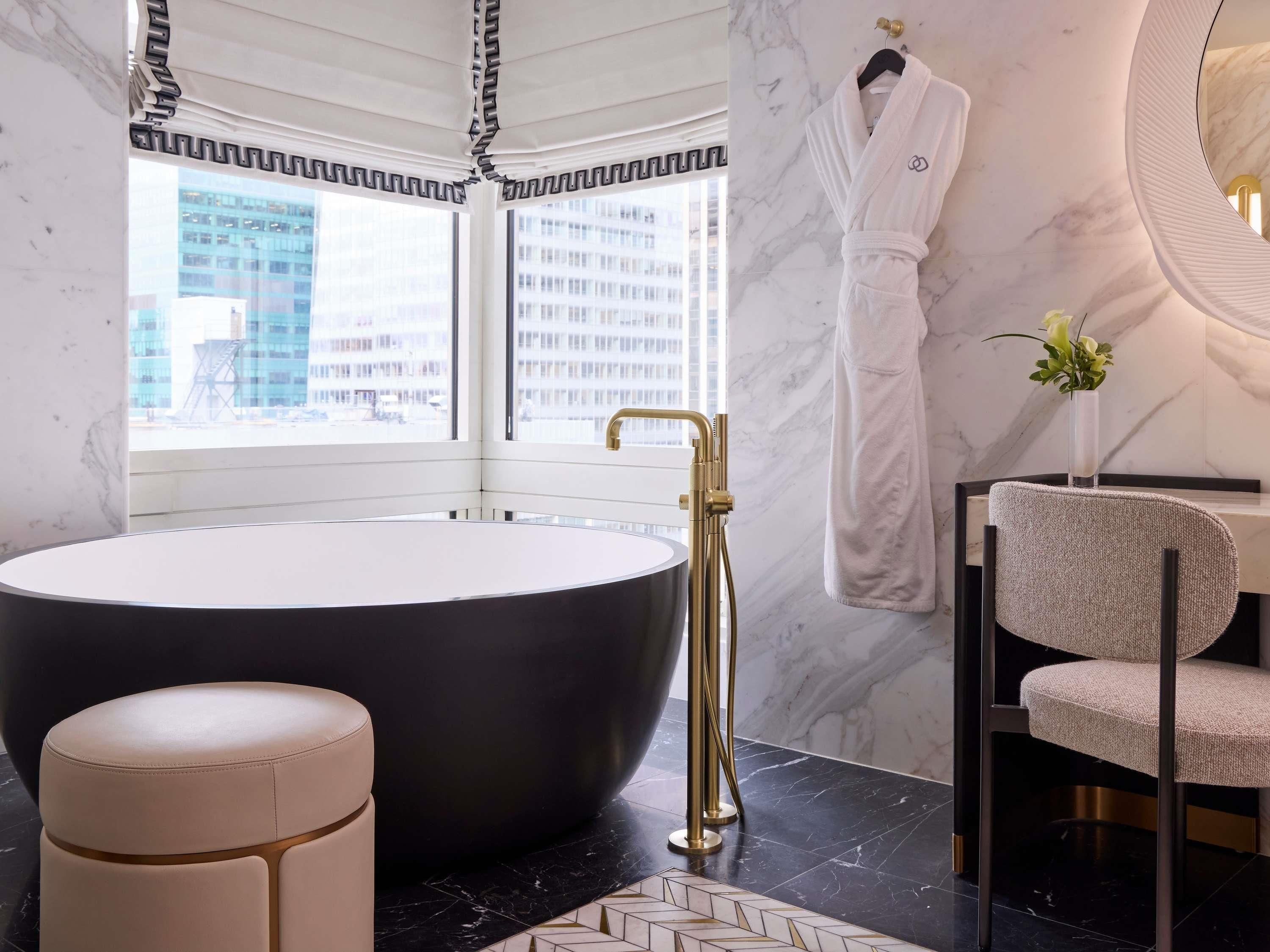 Une salle de bain moderne avec une grande baignoire, un robinet doré, un peignoir blanc accroché au mur et une vue sur la ville à travers de grandes fenêtres.
