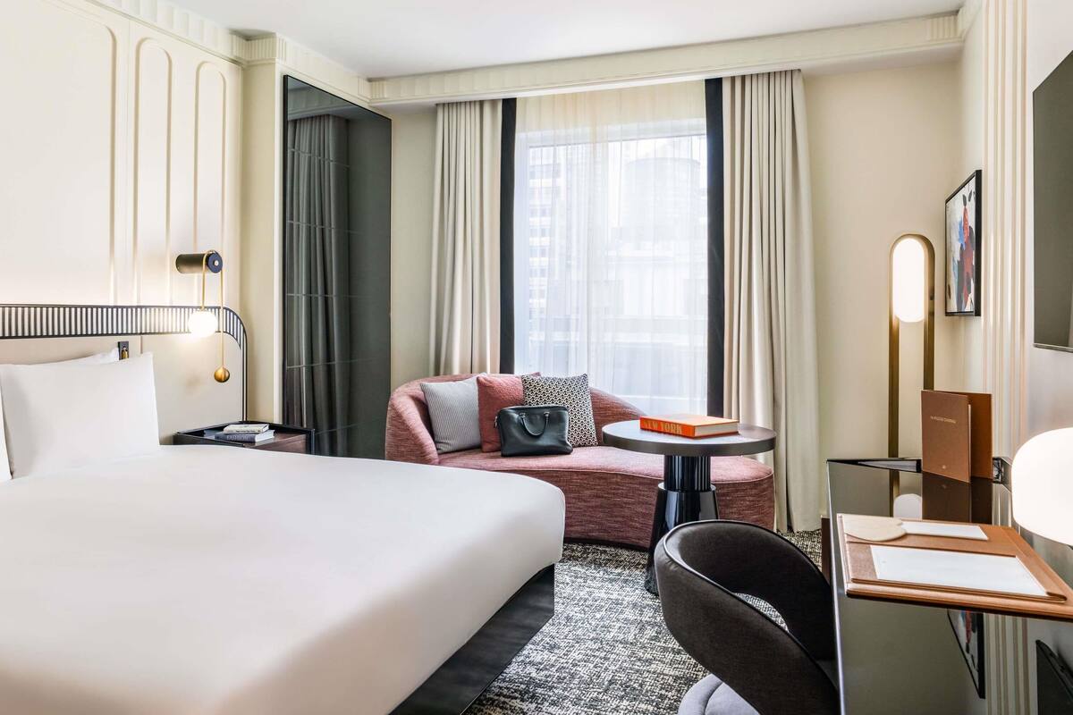 A hotel room with a large bed, a desk with a chair, a sofa, and a window with curtains.