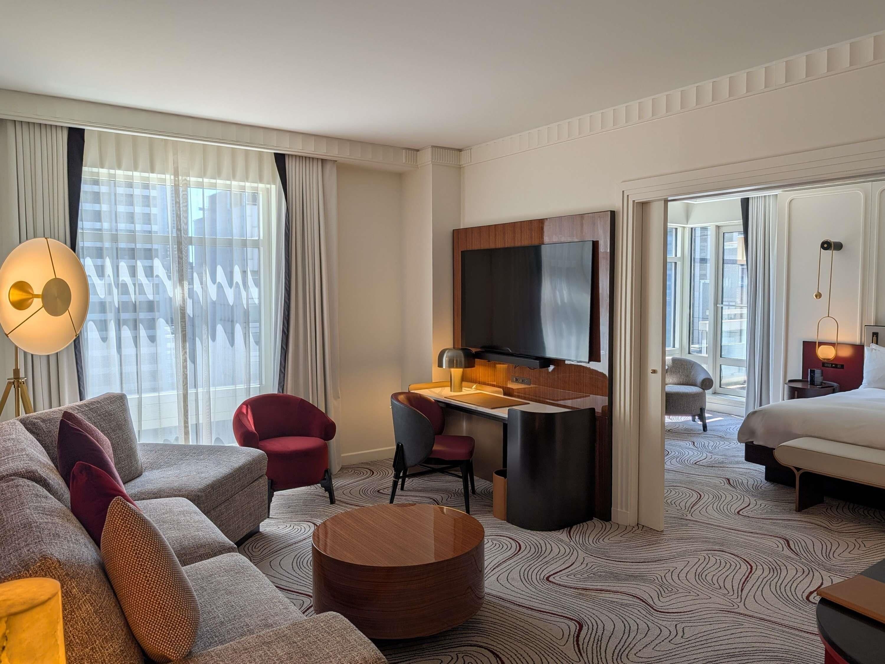 A hotel room with a sofa, a desk, a TV, a bed, and a view of the city.