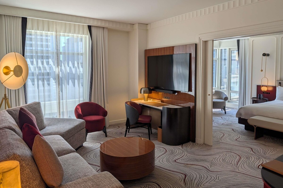 A hotel room with a sofa, a desk, a TV, a bed, and a view of the city.
