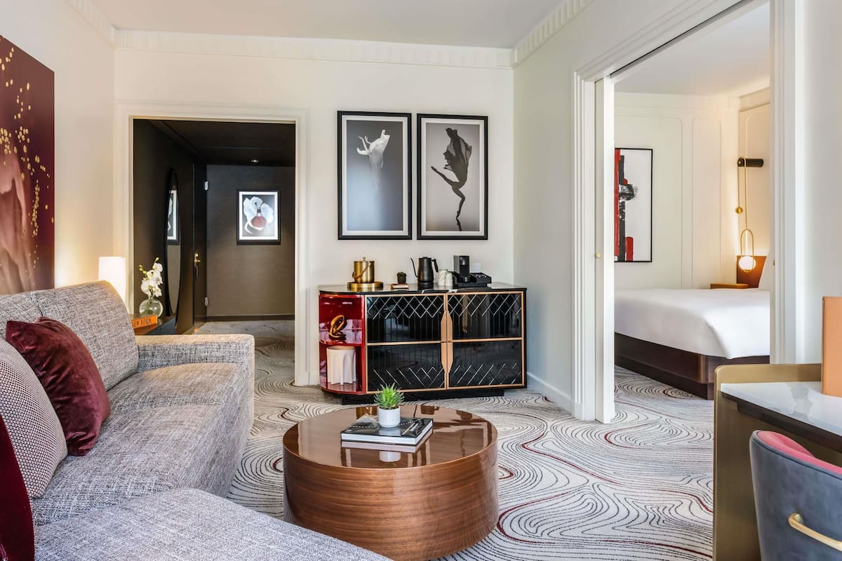 A modern hotel room with a sofa, a round coffee table, a side table with a lamp, a bed, and a console table with decorative items.