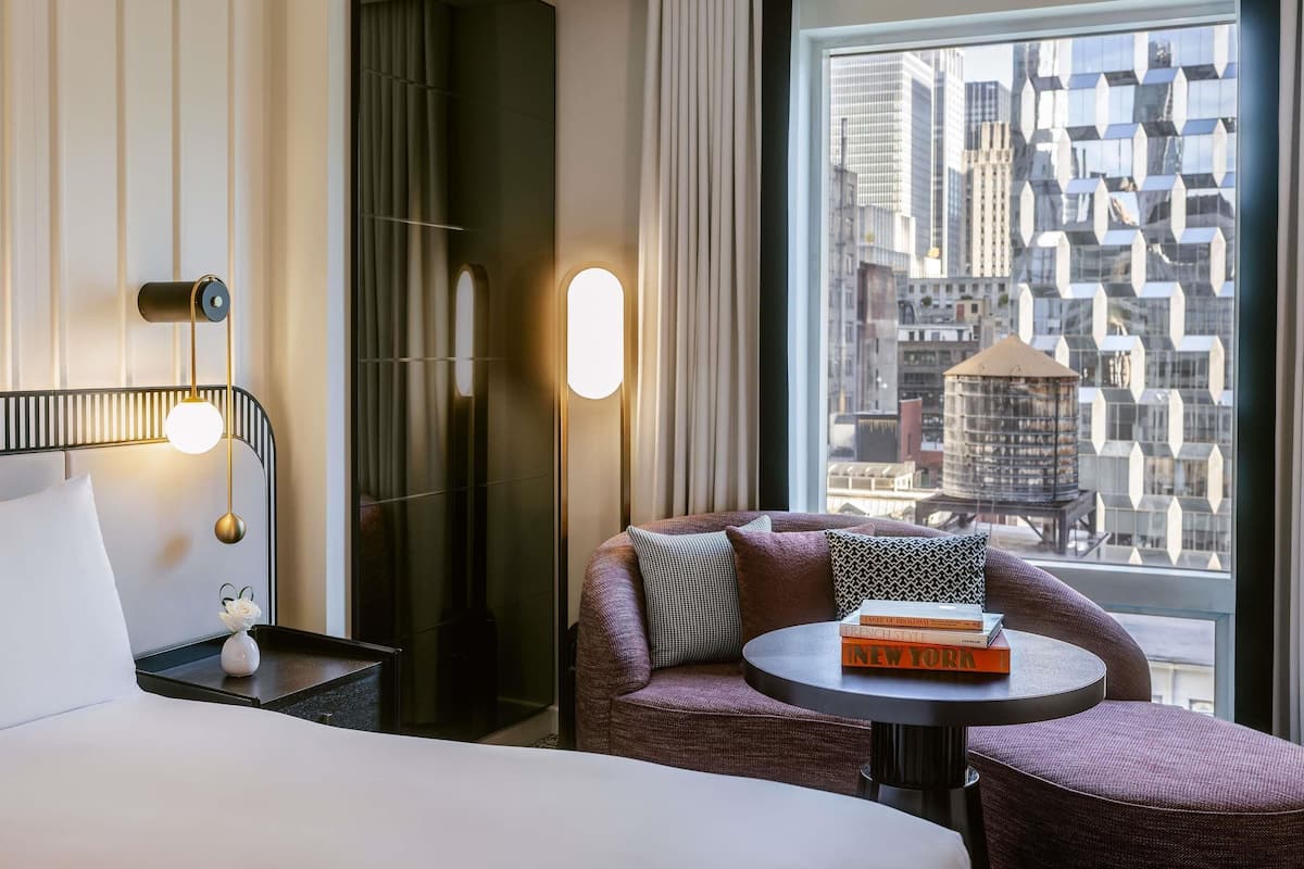 A hotel room with a bed, a round table with books, a chair, a wall-mounted lamp, and a large window with a city view.