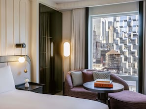 A hotel room with a bed, a round table with books, a chair, a wall-mounted lamp, and a large window with a city view.