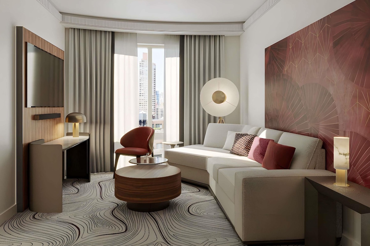 A modern hotel room with a sofa, armchair, round coffee table, and a large abstract painting.