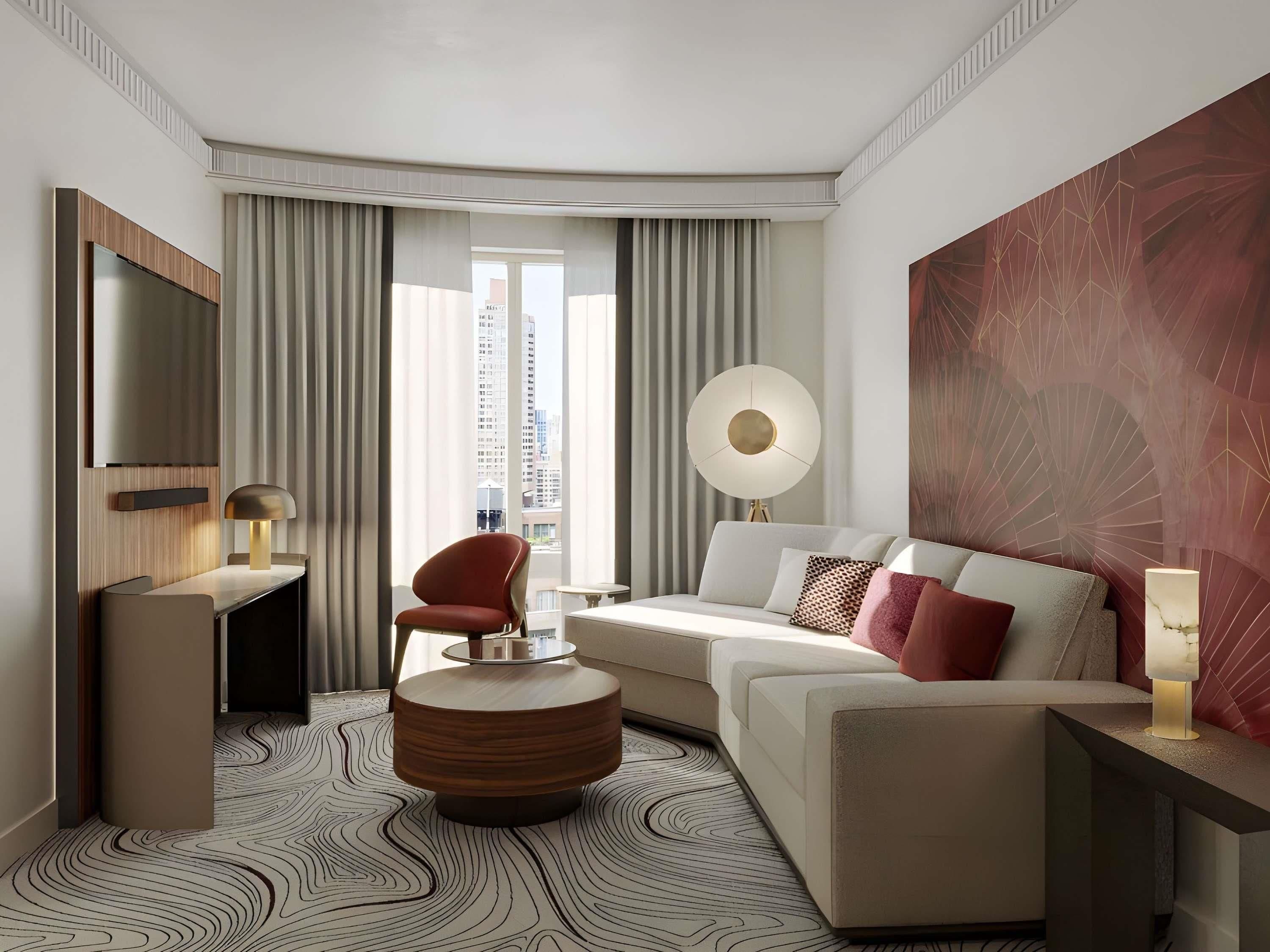 A modern hotel room with a large window, a round wooden coffee table, a sofa with multiple pillows, a side table with a lamp, and a large abstract painting on the wall.