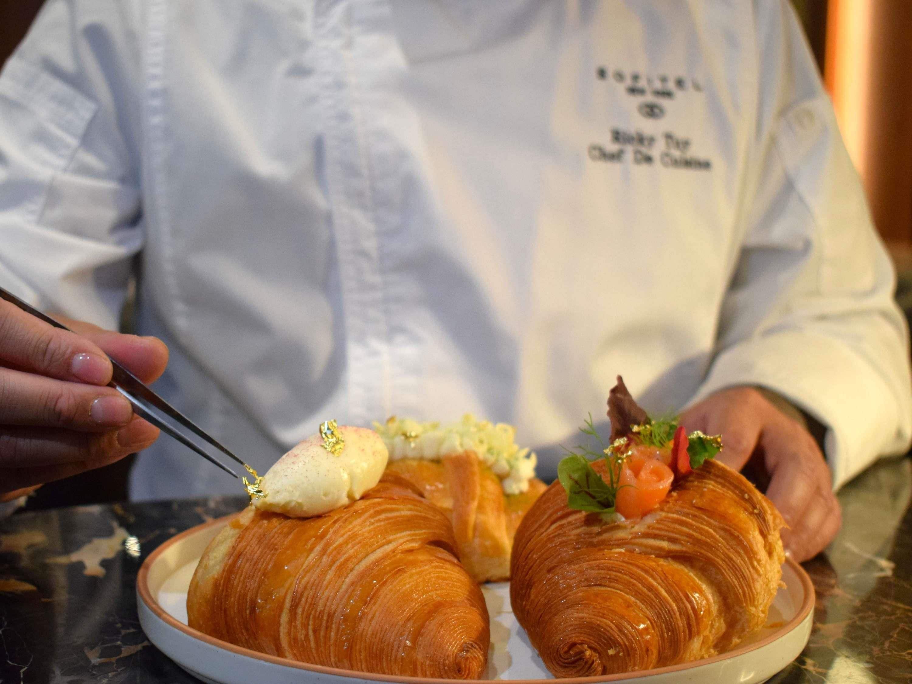 A chef in a white uniform with a name tag, holding a plate with two croissants and a spoon with a dollop of cream.
