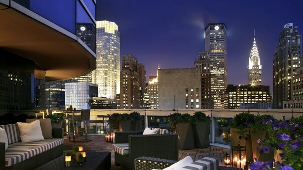 A rooftop terrace with outdoor seating, city skyline views, and potted plants.