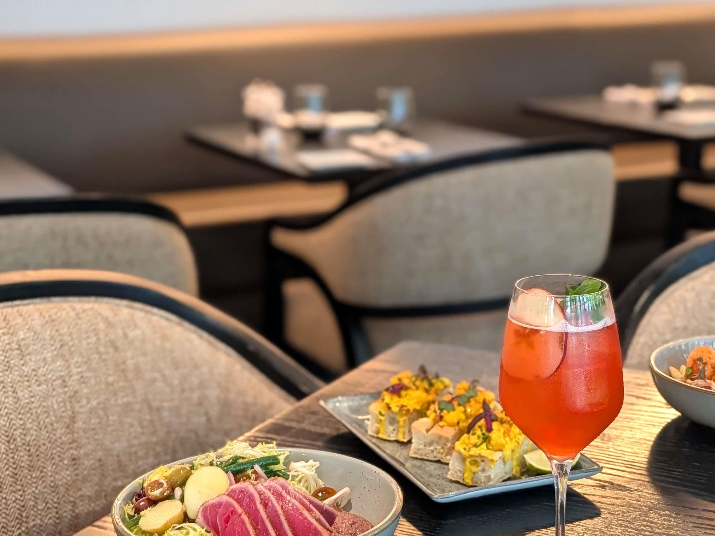 Ein Essbereich mit Tischen, Stühlen und verschiedenen Speisen, darunter eine Salatschüssel, eine Vorspeisenplatte und ein Glas rotes Getränk.