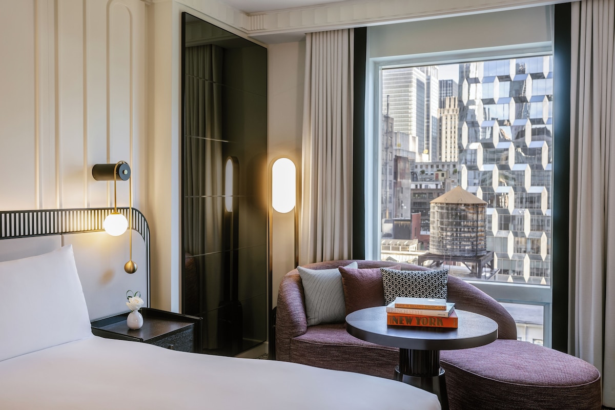 Habitación de hotel con cama, mesita de noche con lámpara, zona de estar con una mesa y un amplio ventanal con vistas a la ciudad.