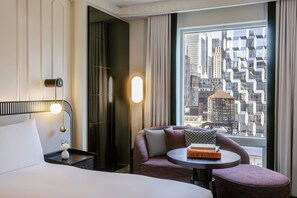 Habitación de hotel con cama, mesita de noche con lámpara, zona de estar con una mesa y un amplio ventanal con vistas a la ciudad.