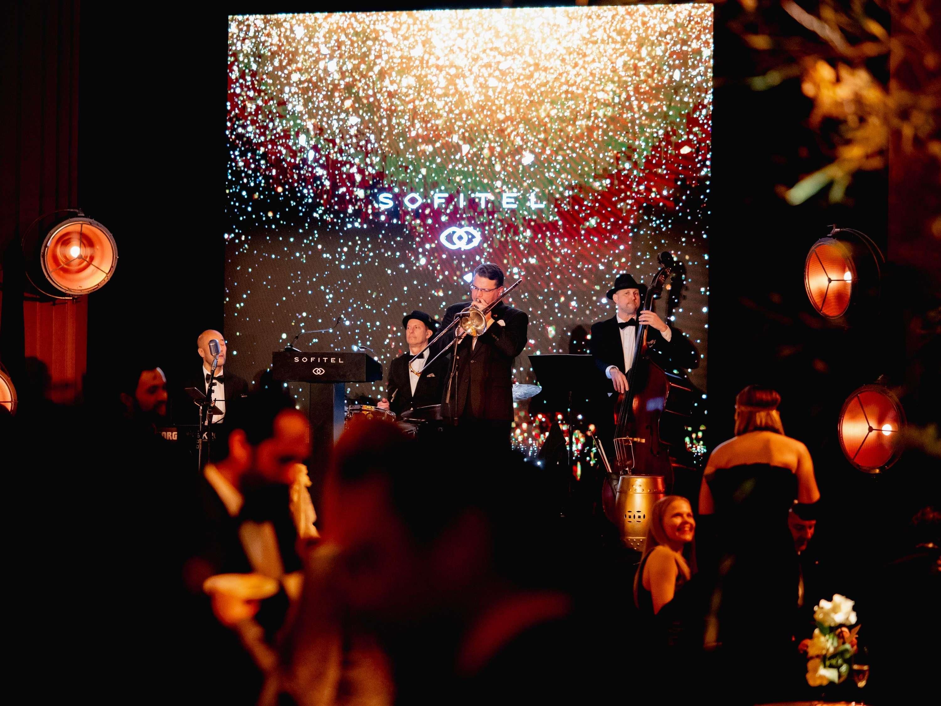 A band performing on a stage with a backdrop of lights and a microphone stand labeled 'SOFITEL'.