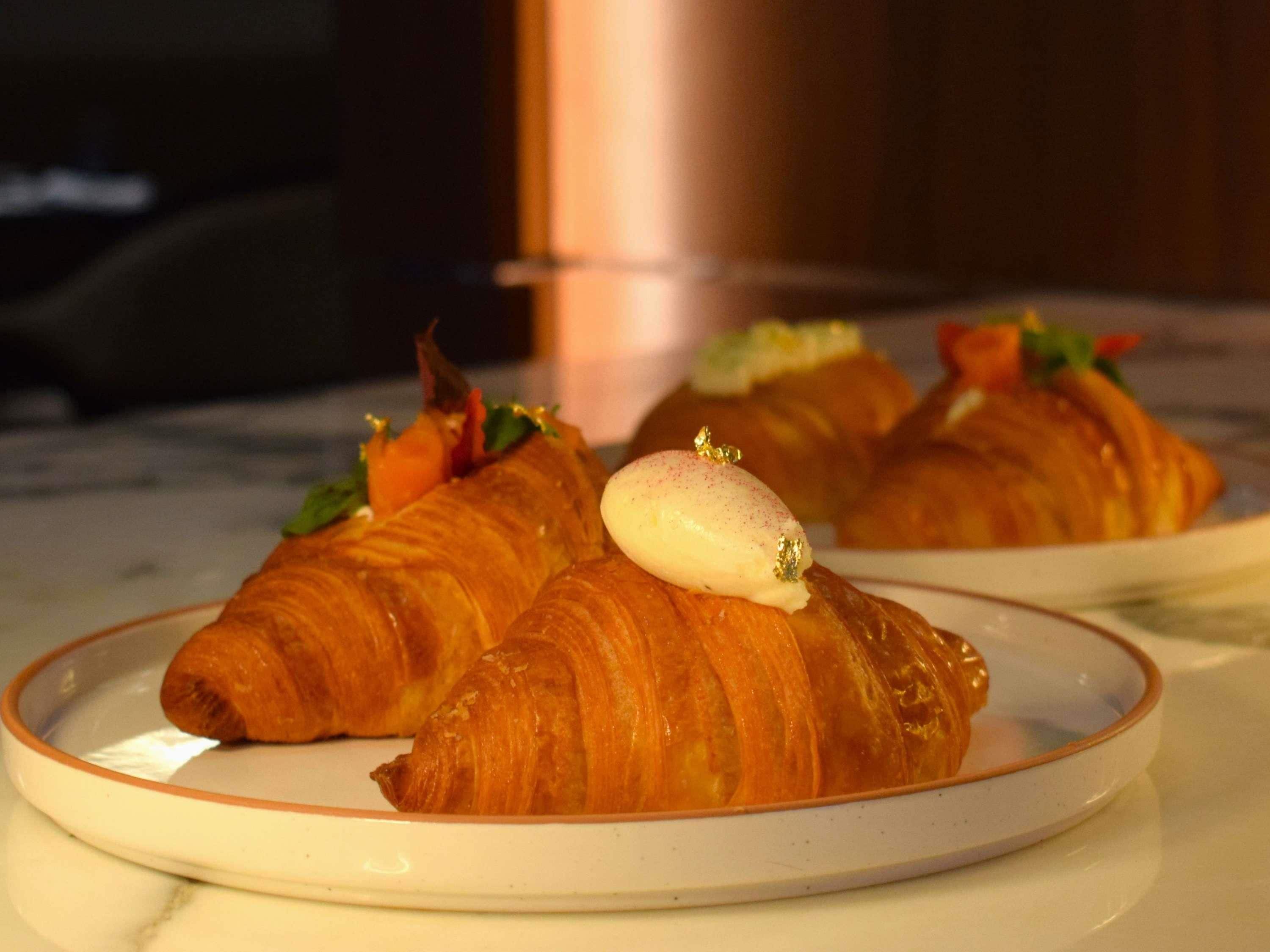Tre croissanter på tallerkener med dessert ovenpå, placeret på et bord.