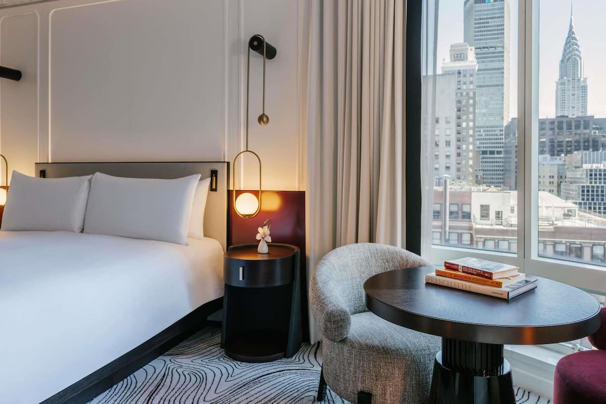 A hotel room with a bed, a round table with books, a chair, a nightstand with a lamp, and a view of the cityscape.