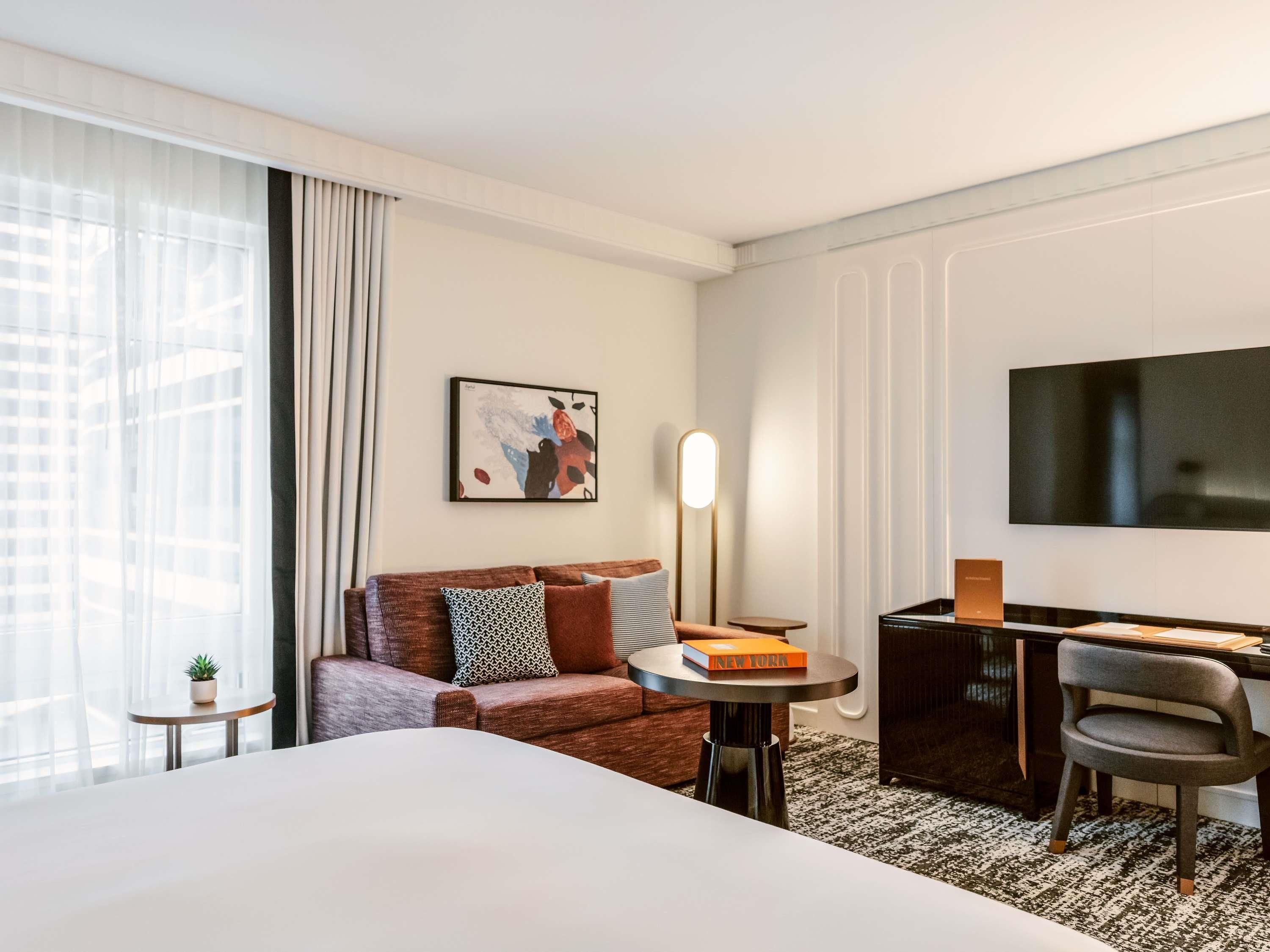 Et moderne hotelværelse med en stor seng, en sofa, et skrivebord med stol, et fjernsyn og et lille bord med en plante.
