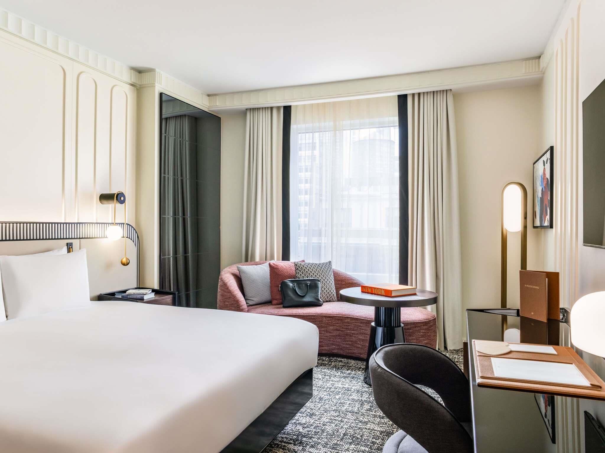 A hotel room with a bed, a desk, a chair, a sofa, and a window with curtains.