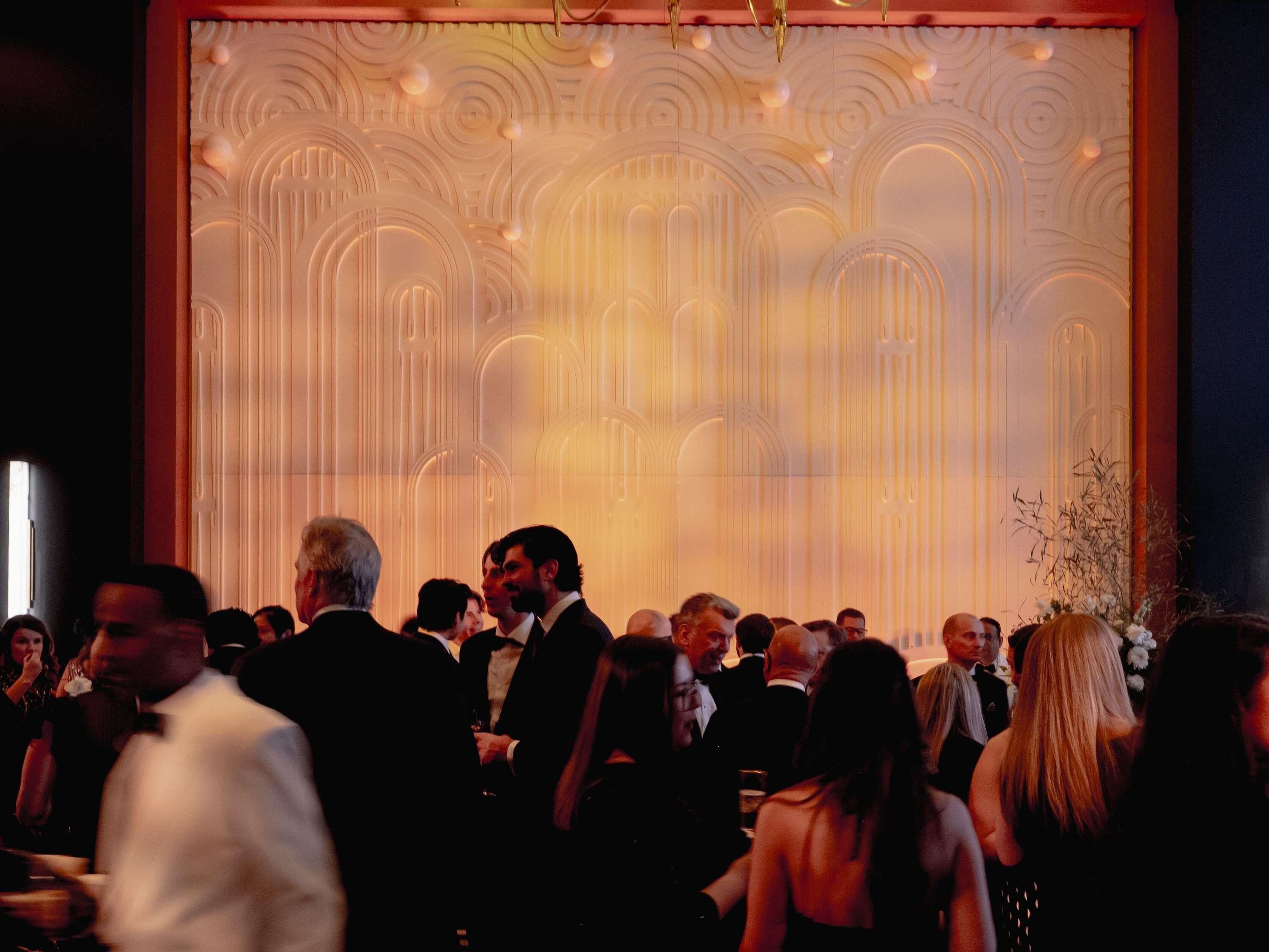 A crowded indoor event with a decorative backdrop featuring arches and vertical lines.