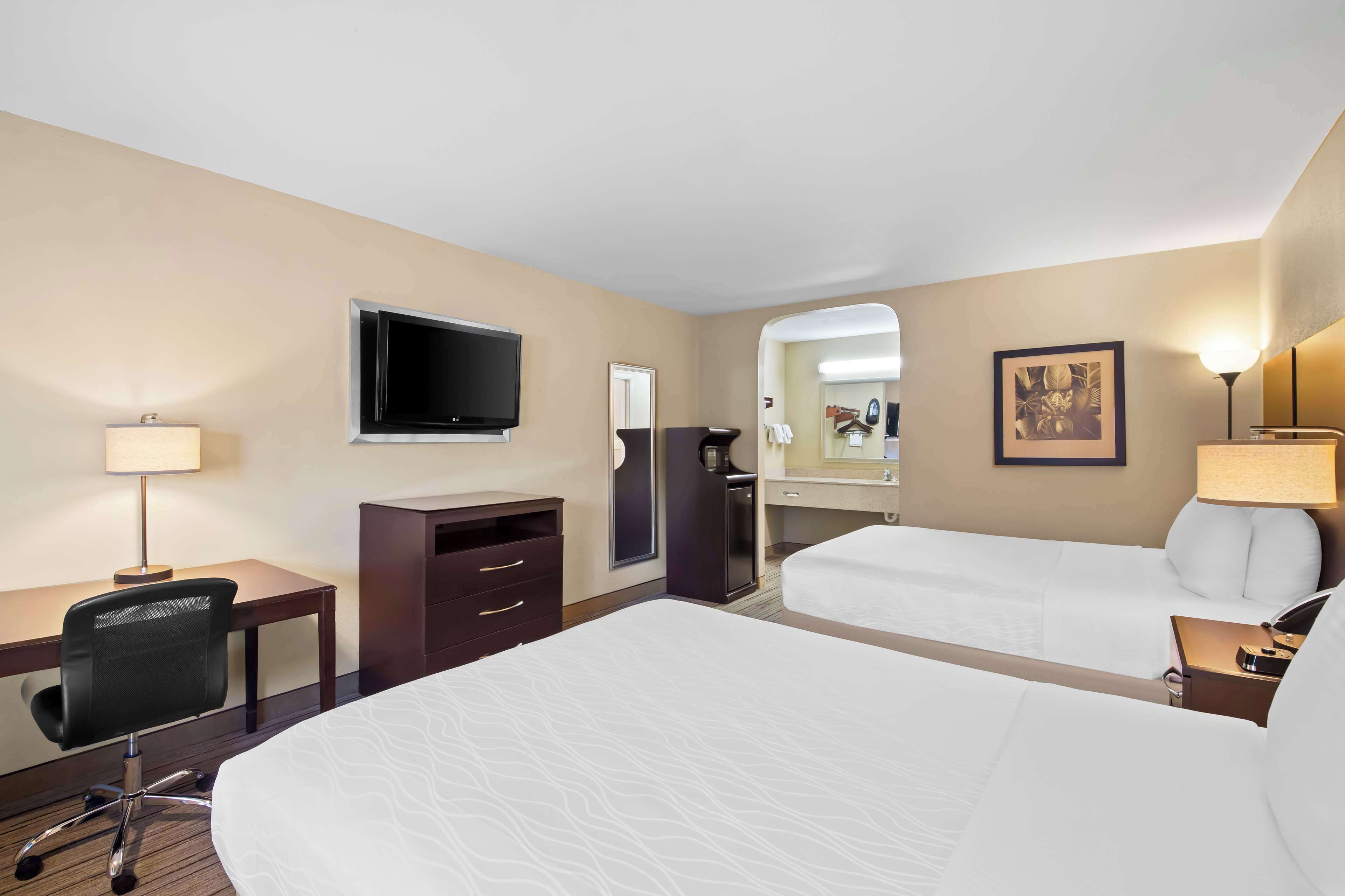 A hotel room with a large bed, a desk with a chair, a television, a desk lamp, a nightstand, and a bedside table.