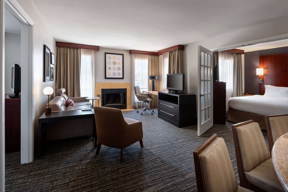 A hotel room with a bed, a desk, a chair, a TV, a fireplace, and a large window with curtains.
