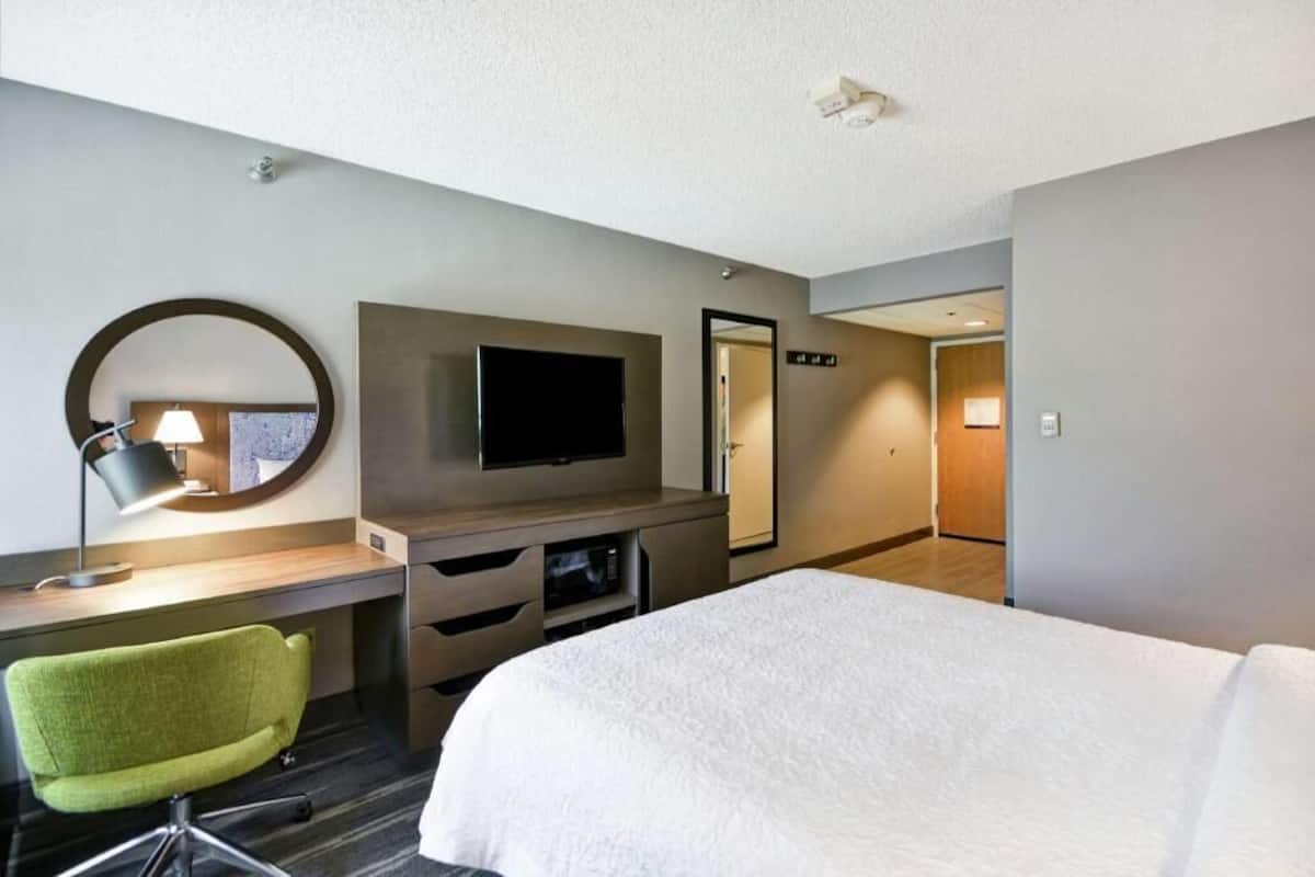 A hotel room with a bed, a desk with a lamp, a TV, a mirror, and a door.