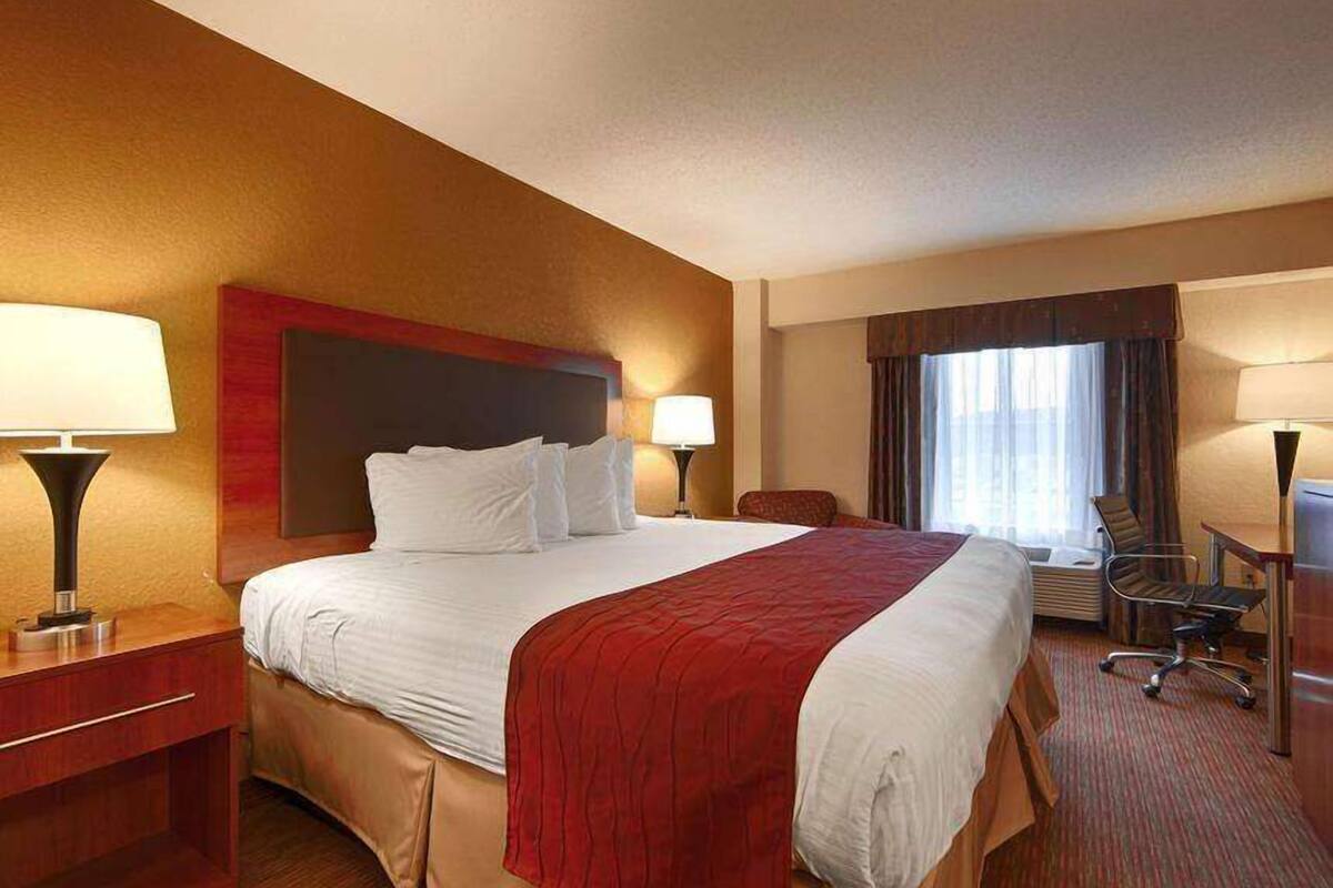 A hotel room with a large bed, two bedside tables with lamps, a desk with a chair, and a window with curtains.