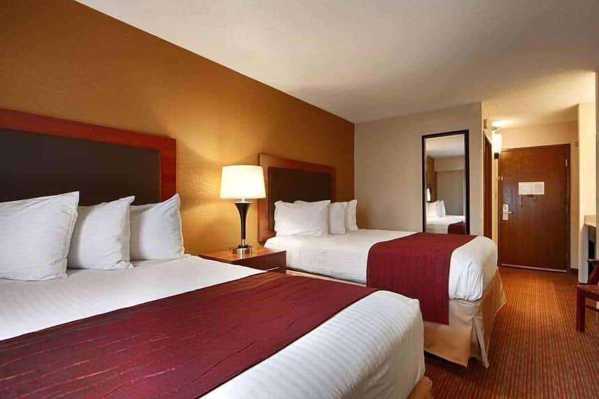 A hotel room with two beds, a nightstand with a lamp, a mirror, and a door.