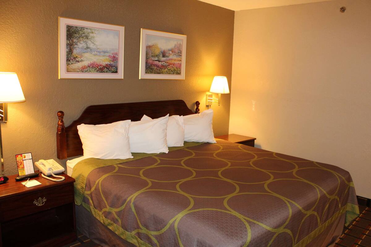 Habitación de hotel con una cama grande, dos cuadros colgados en la pared, una mesita de noche con un teléfono y una lámpara, y una lámpara de pared.