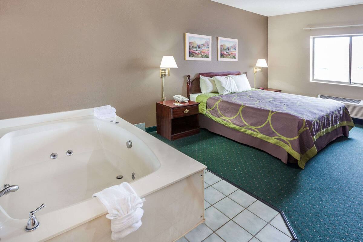 Una habitación de hotel con una bañera grande, una cama con cubrecama verde y morado, dos lámparas de pared y dos cuadros en la pared.