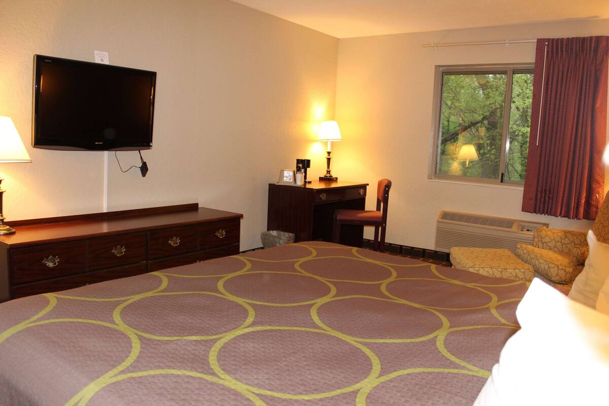 Habitación de hotel con cama, escritorio con lámpara, televisor montado en la pared y ventana con cortinas.