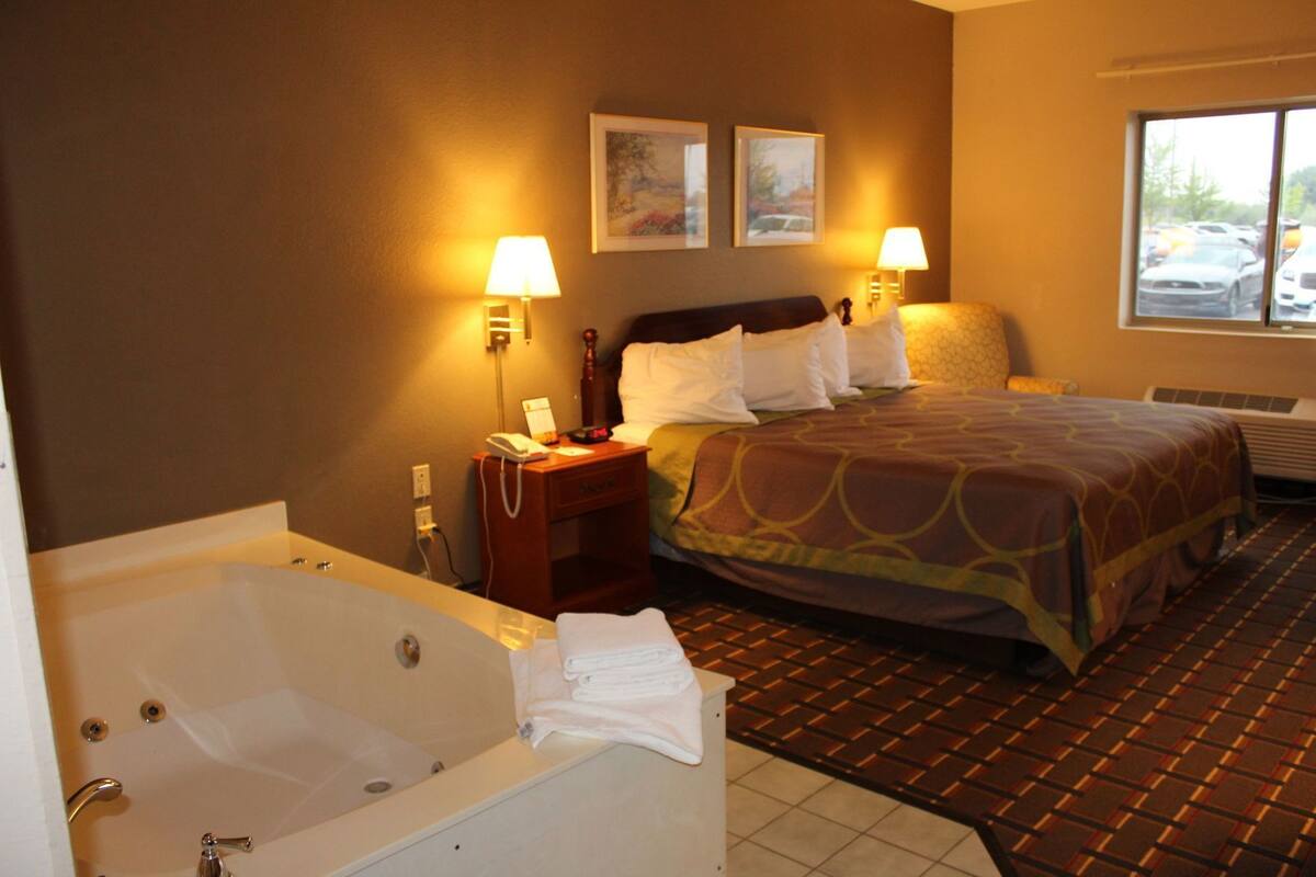 Habitación de hotel con una bañera grande, una cama con cubrecama estampado, dos lámparas de pared y una ventana con vista al estacionamiento.